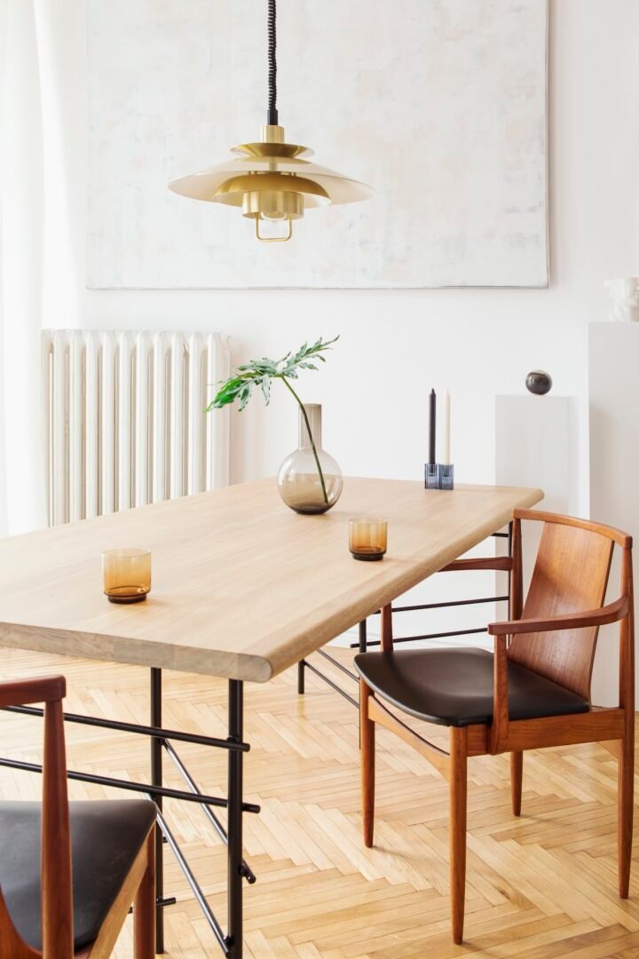 Stylish and modern dining room interior with design sharing table, chairs, gold pendant lamp, abstract paintings and elegant accessories. Tropical leafs in vase. Eclectic home decor.