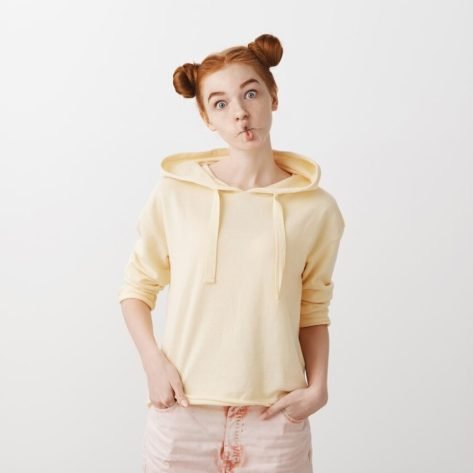 Studio shot of cute stylish young redhead with two buns making fish face with folded lips, popping eyes at camera, mimicking and making fun, standing childish against gray background. Emotions concept